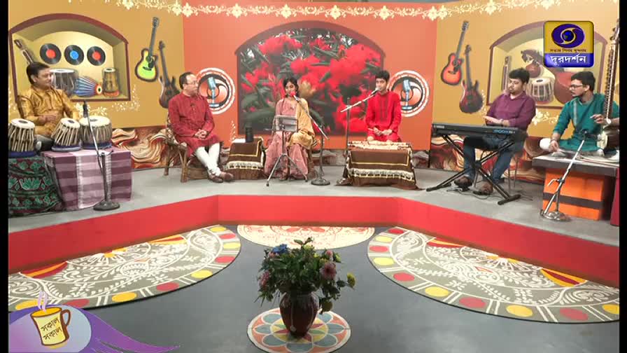 A musical ensemble, likely for a DD Bangla broadcast, is arranged on a stage with various instruments, including tabla drums and a keyboard. The musicians, dressed in traditional Indian attire, are seated and ready to perform.
