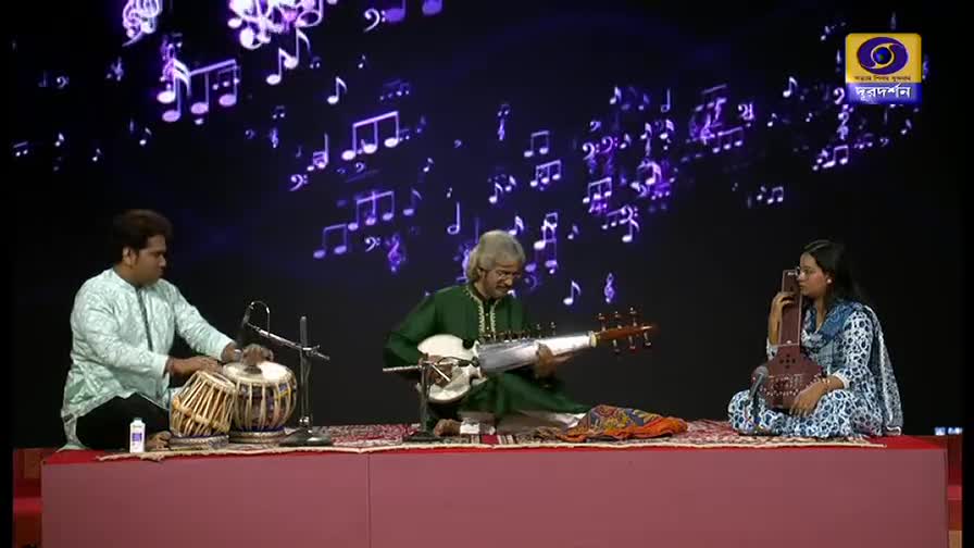The man in green, with his long instrument, is plucking the strings. Beside him, the tabla player's hands move quickly, and the woman holds her instrument, ready to play.

