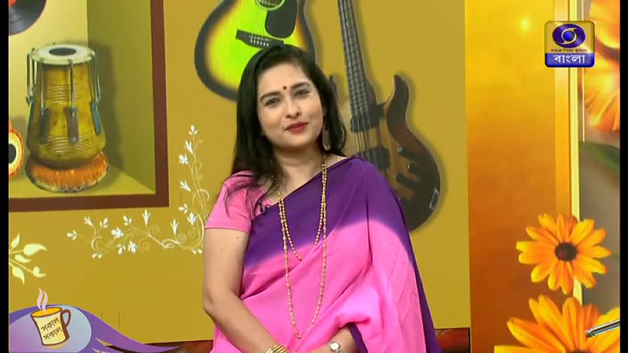 A woman in a pink and purple saree smiles at the camera, a long necklace draped around her neck. The DD Bangla logo sits in the corner, and a tabla drum and guitars are visible in the background.
