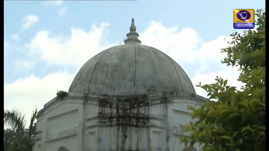 A large, domed structure dominates the frame, its white surface weathered with dark streaks. The logo for DD Bangla is visible in the corner, and the sky is a pale blue.
