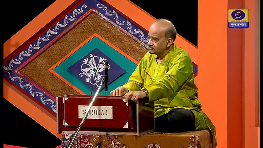 A man in a bright green shirt sits playing a harmonium, his hands moving over the keys. Behind him, a colorful geometric backdrop and the DD Bangla logo mark the stage.

