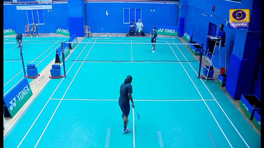 A badminton match is underway, with players positioned on opposite sides of the court, ready to serve. The DD Bangla logo is visible in the corner, broadcasting the game.
