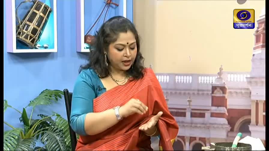 The woman in the red sari gestures with her hands while speaking, a small item resting in her palm. Behind her, a framed logo and a backdrop of a building suggest a DD Bangla studio.
