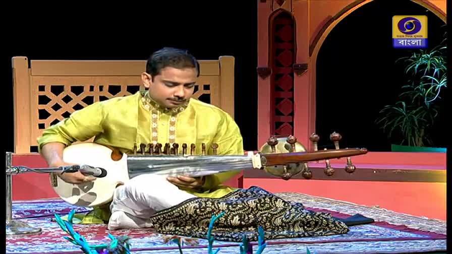 A musician in a lime green kurta sits cross-legged, gently plucking the strings of a sarod. The DD Bangla logo is visible as he plays, the stage lit with warm colors.
