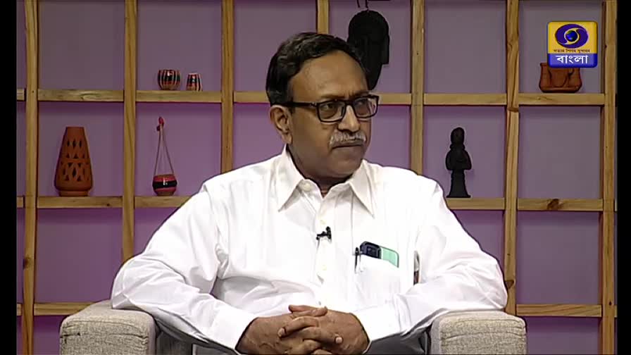 A man in a white shirt sits in a chair, looking off-camera, with his hands clasped. Behind him, a wooden shelf holds various decorative items, and the DD Bangla logo is visible in the corner.
