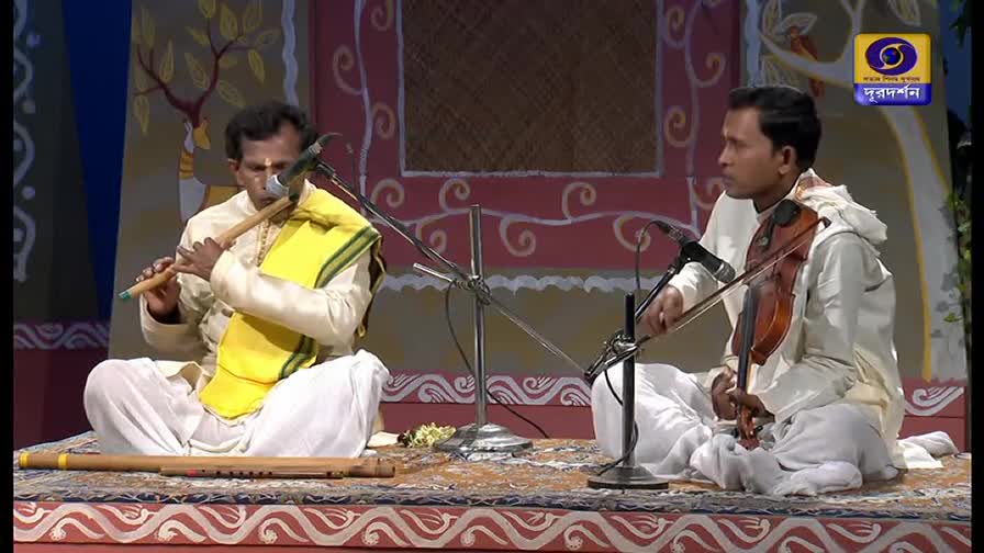 A man in a yellow shawl blows into a flute, while another plays a violin, both seated on a patterned rug. Microphones are positioned in front of them, likely for a performance on DD Bangla.
