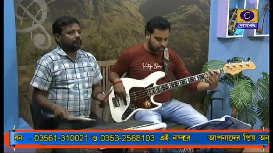 A man in a checkered shirt taps a drum pad with two sticks while another strums a white bass guitar.  The backdrop is a simple studio set, with a DD Bangla logo visible.

