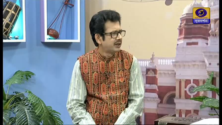 The man with glasses, wearing a patterned vest, is speaking animatedly. He's on a set, with the DD Bangla logo visible on the right.
