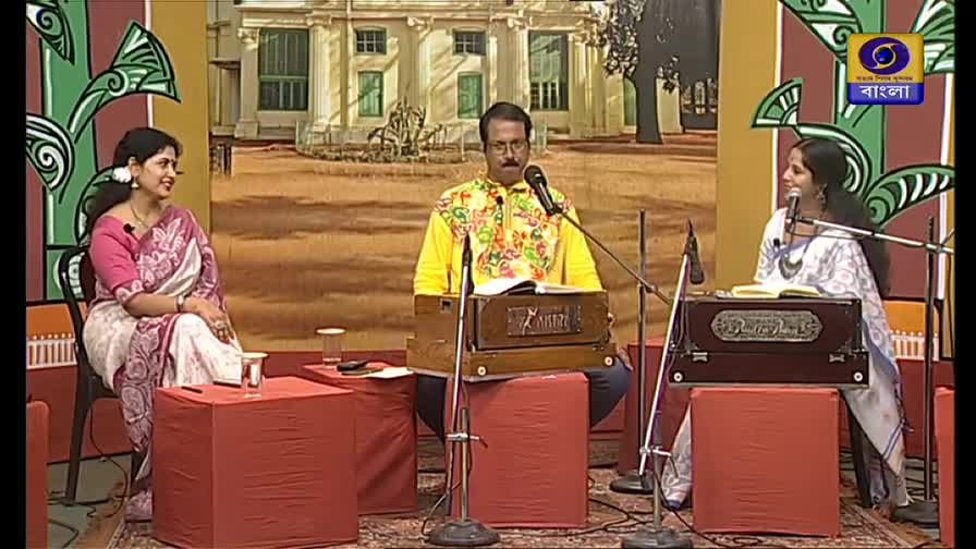 Three musicians sit on stage, each facing a harmonium and a microphone. The man in the center, wearing a bright yellow shirt, appears to be leading the performance on this DD Bangla program.
