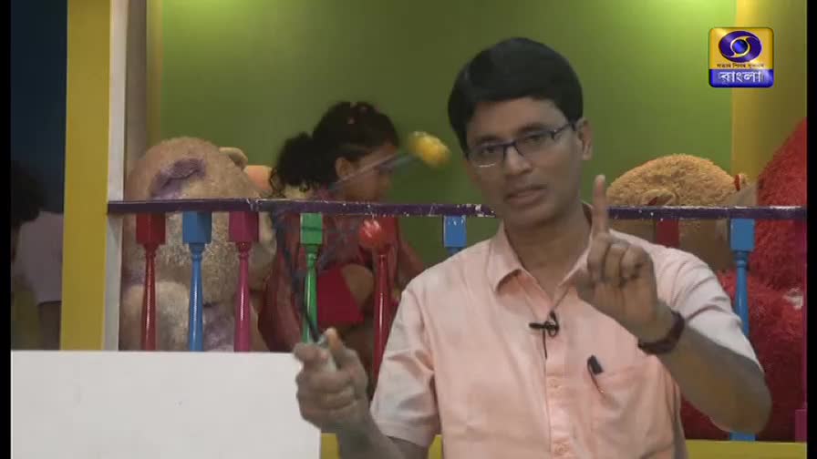 A man in glasses gestures with his fingers, as if explaining something. Behind him, a child is playing in a colorful play area, visible on DD Bangla.
