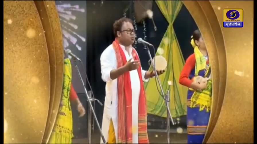 The man in the center, wearing a red and white scarf, sings into a microphone while holding a small, round instrument.  Two other performers stand nearby, dressed in colorful traditional clothing, also singing.
