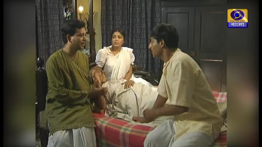 A man in a green kurta gestures emphatically, talking to another man seated on a bed. A woman in a white sari sits beside a person lying down, watching the exchange.
