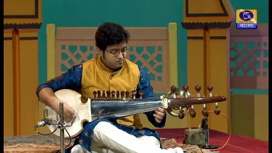 A musician, wearing a yellow vest and blue sleeves, sits cross-legged and plays a stringed instrument. The logo for DD Bangla is visible in the corner.
