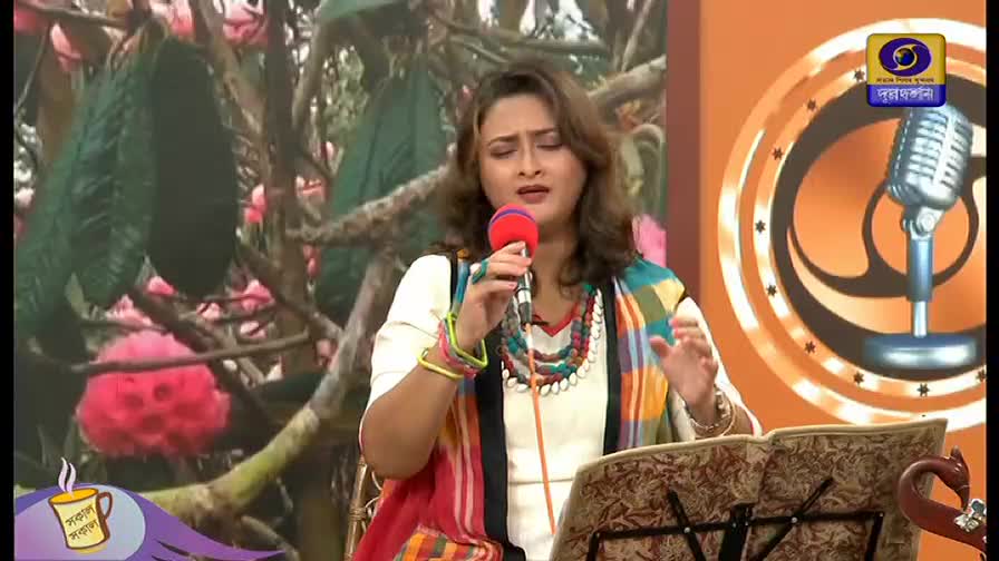 A woman, dressed in a white top and colorful shawl, sings into a red microphone, her hands moving to the music. Behind her, the DD Bangla logo and a backdrop of flowering branches suggest a studio performance.
