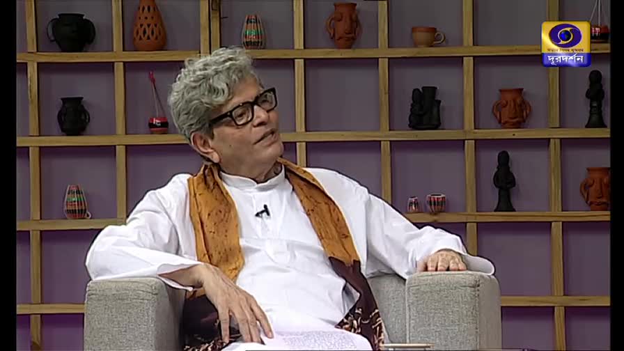 A man in glasses and a white kurta speaks, gesturing with his hands as he sits in a studio chair. Behind him, a shelf displays various pottery pieces, and the DD Bangla logo is visible in the corner.
