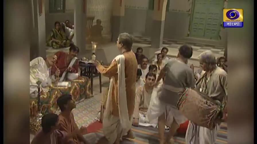 A man in a saffron robe holds a lit torch, gesturing towards a group of seated people. Two men are playing a drum as they move in a circle, while others watch from the floor and a raised platform.
