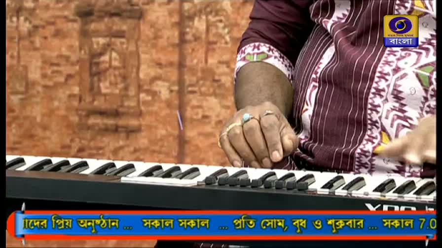 A person's hands are poised over a keyboard, fingers ready to play. The DD Bangla logo is visible, and the text below mentions "Sokal Sokal" and the days of the week.
