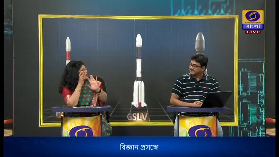 On the DD Bangla set, a woman gestures animatedly while speaking, her hands wide. Across from her, a man in a striped shirt looks on, a laptop open on his desk.
