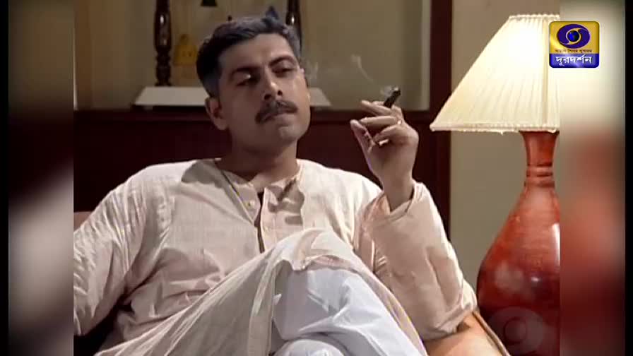 A man, dressed in simple clothes, sits and smokes a cigar, the smoke curling upwards. A DD Bangla logo is visible in the corner, hinting at the program's origin.
