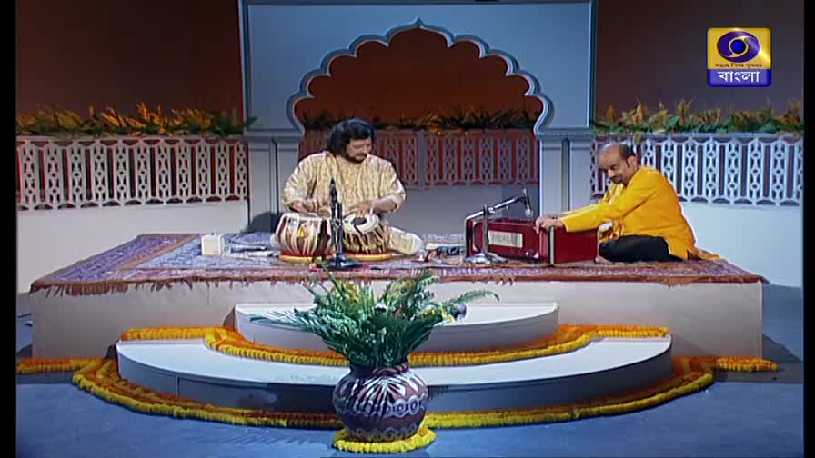 A man in a cream-colored kurta plays the tabla, his hands a blur above the drums. Beside him, another man in a yellow shirt works the bellows of a harmonium, the music broadcast on DD Bangla.
