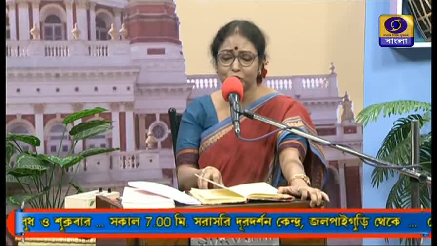 A woman in a sari speaks into a microphone, looking down at a notebook on the desk in front of her. The DD Bangla logo is visible, and the screen shows text in Bengali at the bottom.
