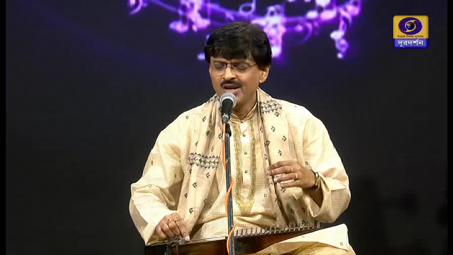 A man in glasses and a patterned shawl sings into a microphone, his fingers gently plucking at a stringed instrument. The DD Bangla logo is visible in the corner, indicating a broadcast from India.

