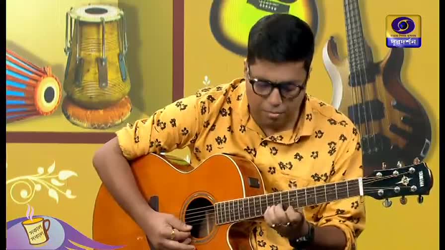 A man in a patterned yellow shirt concentrates on playing an acoustic guitar. The DD Bangla logo is visible on the right, and a tabla sits in the background.
