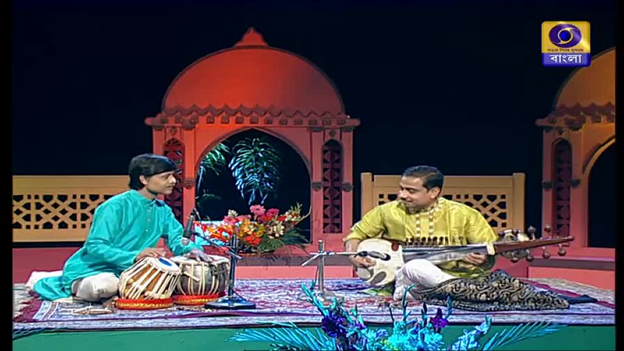 On a DD Bangla set, a young man in a teal kurta taps the tabla drums, while another man in a yellow shirt plucks the strings of a sarod. The musicians are seated on a patterned rug, with a decorative archway behind them.
