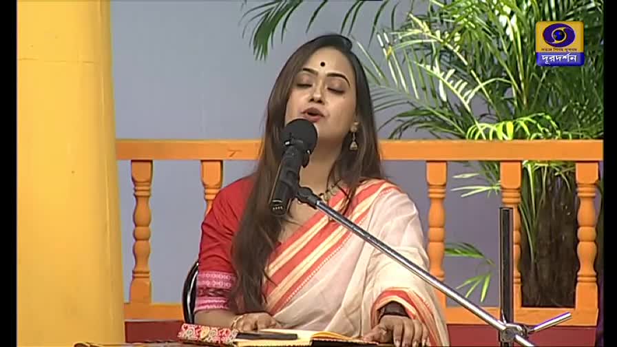 A woman in a red and cream sari sings into a microphone on a stage, her eyes closed. Behind her, the DD Bangla logo is visible, framed by a backdrop of greenery and an orange railing.
