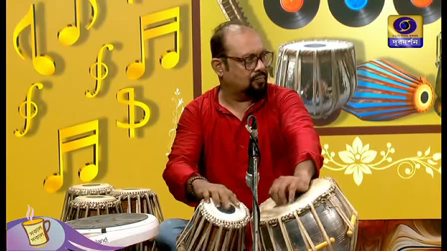 The man in the red shirt is seated, his hands moving deftly over the tabla drums. A DD Bangla logo sits in the corner, and the set is decorated with musical notes.
