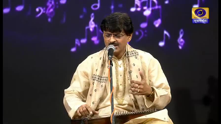 The musician, wearing glasses and a patterned scarf, sings into a microphone. His hands move over the strings of a stringed instrument, likely a *santoor*, as the music plays on DD Bangla.
