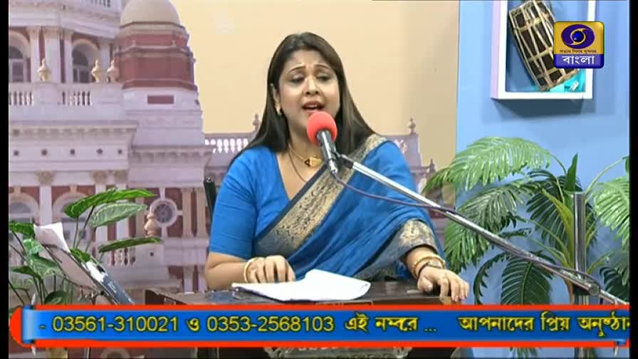 A woman in a blue sari speaks into a red microphone, her expression animated. Behind her, a backdrop depicts a building, and the DD Bangla logo is visible on the wall.
