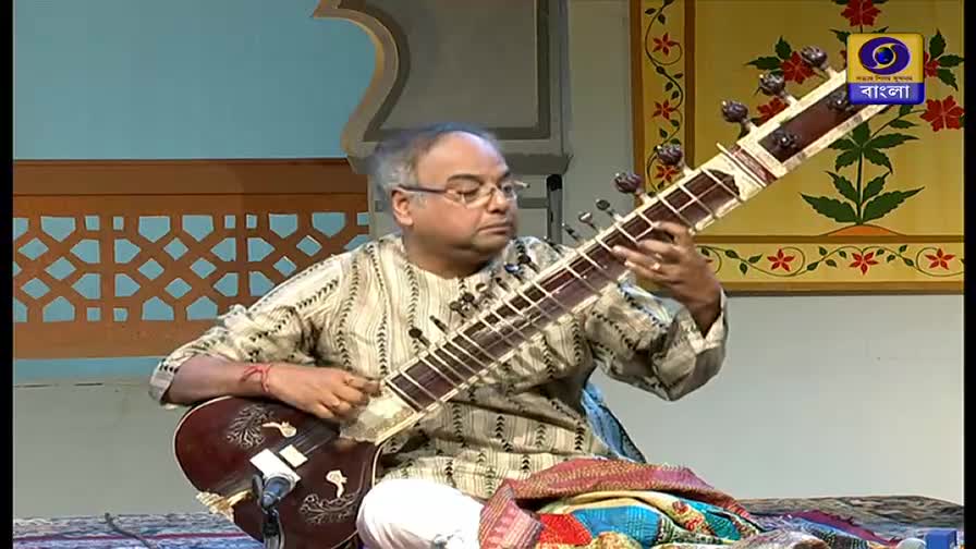 The man, seated, plucks the strings of a long-necked sitar, his fingers dancing across the frets. A DD Bangla logo is visible in the background, as the musician plays in a studio setting.
