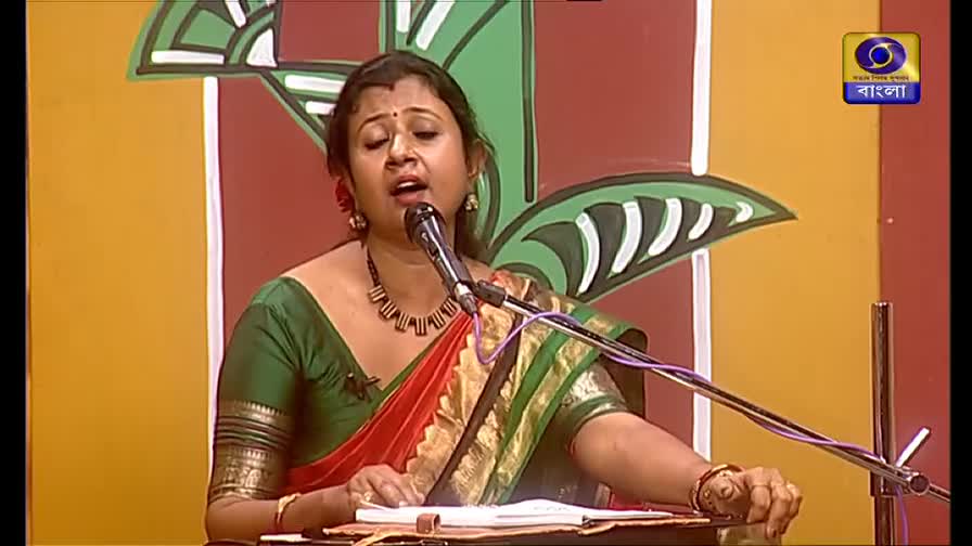 A woman in a green blouse and red sari sings into a microphone, eyes closed. The DD Bangla logo sits in the corner as she performs on a brightly colored set.

