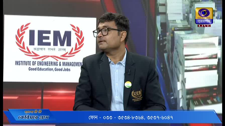 A man in a blazer and glasses is speaking on a DD Bangla program, with the IEM logo behind him. The screen displays a phone number at the bottom.
