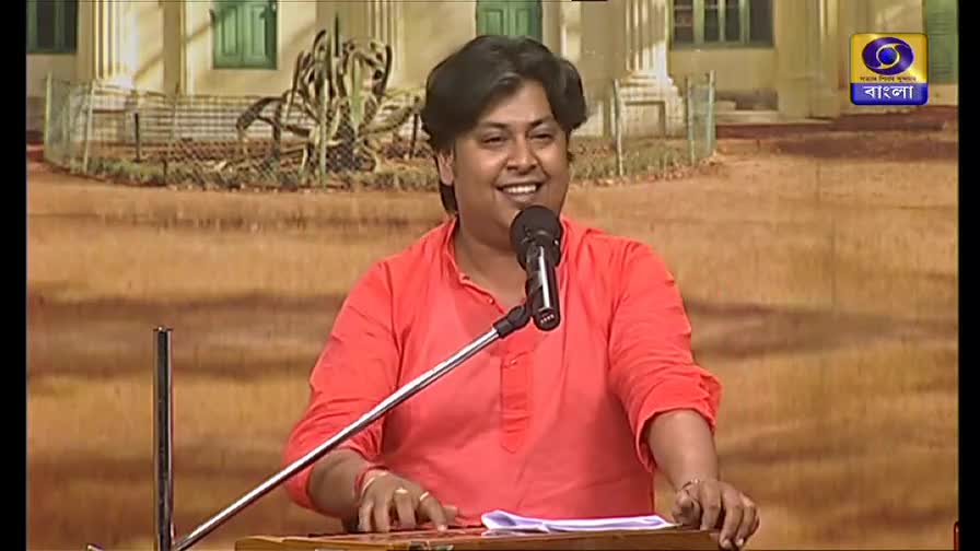 The man in the red shirt smiles into the microphone, his fingers dancing across the keys of a harmonium. Behind him, a backdrop suggests a garden setting, and the DD Bangla logo is visible in the corner.
