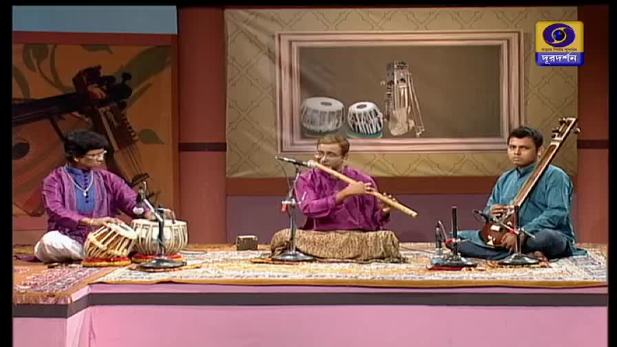 The tabla player's hands are a blur, striking the drums in a steady rhythm. A flautist and a stringed instrument player sit cross-legged, accompanying the percussion on the DD Bangla stage.
