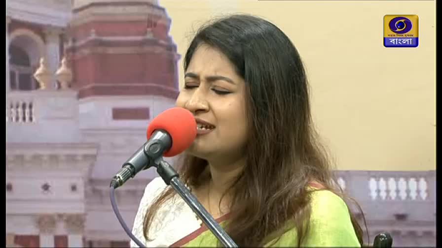 A woman sings into a microphone, her eyes closed, with a backdrop of a building. The DD Bangla logo is in the corner.
