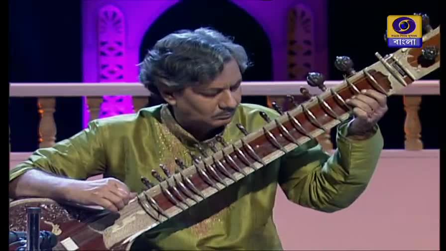 The musician, on what looks like a DD Bangla broadcast, is focused on the sitar, his fingers dancing across the frets. The instrument's long neck extends toward the camera as he plucks the strings.

