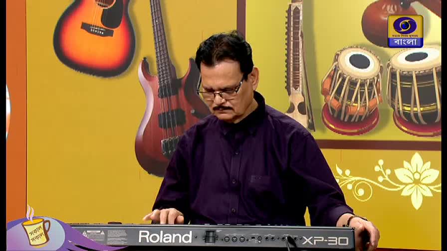 A man in a purple shirt plays a Roland keyboard, his fingers dancing across the keys. Behind him, a DD Bangla logo and various instruments decorate the set.

