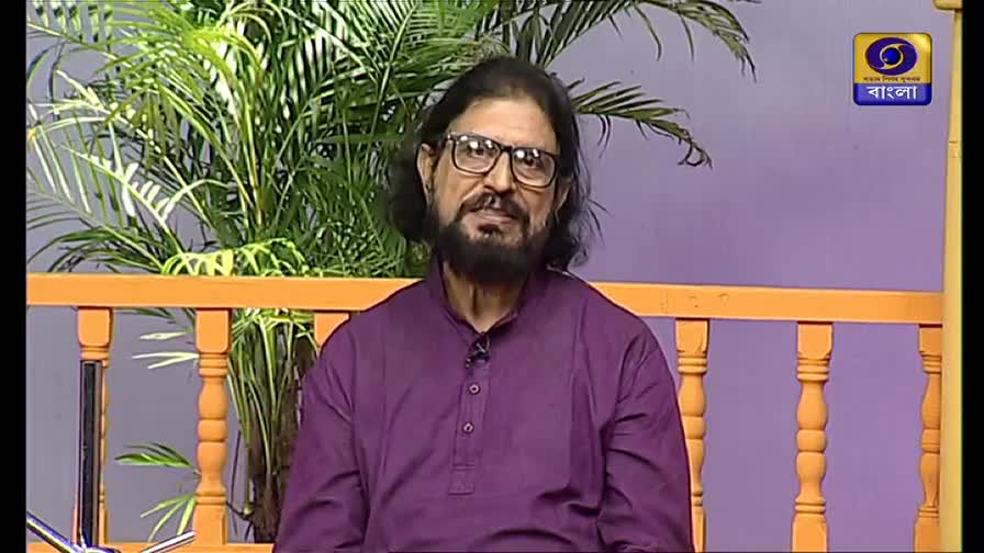 A man with a long beard and glasses sits in front of a plant, speaking on DD Bangla. The logo of the Indian television channel is visible in the corner.
