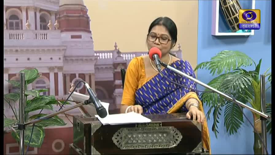 A woman in glasses sings into a microphone, her voice amplified by the equipment. She’s wearing a yellow blouse under a blue patterned sari, and the DD Bangla logo is visible in the corner.
