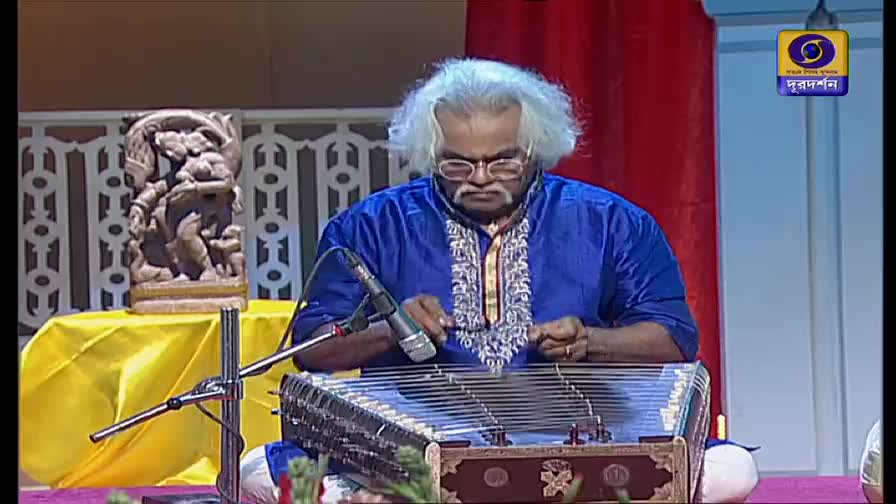 The musician, wearing a blue kurta, gently strikes the strings of his hammered dulcimer. A microphone is positioned close, ready to capture the music for DD Bangla.
