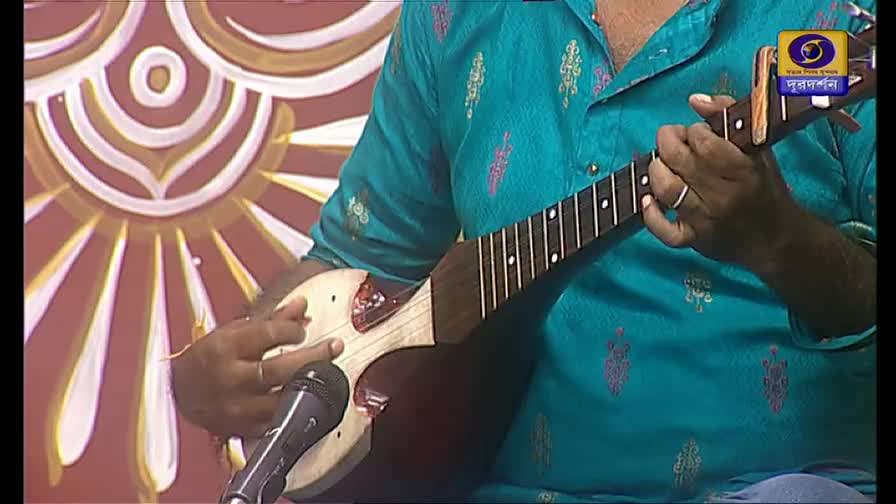 A musician in a teal shirt strums a stringed instrument, his fingers dancing along the fretboard. The logo for DD Bangla is visible in the corner, and a microphone sits nearby.
