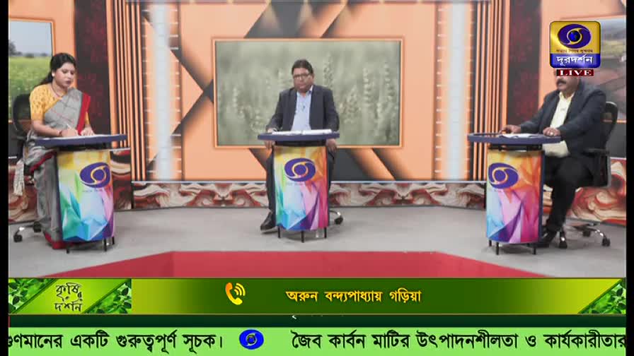 Three people sit at desks on a DD Bangla set, each with a microphone and a colorful stand. The middle man gestures with his hands as he speaks, while the woman on the left takes notes.
