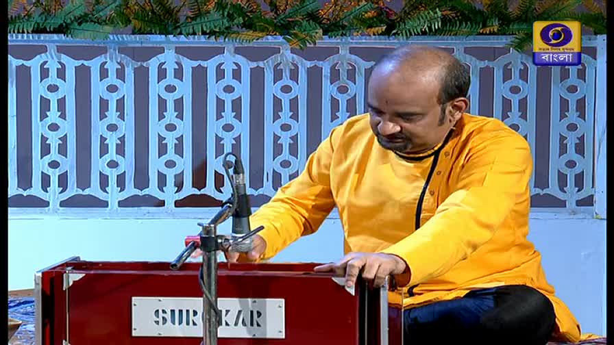 A man in a bright yellow shirt sits playing a red harmonium, his hands moving over the keys. The DD Bangla logo is visible in the corner, and the backdrop suggests a studio setting.
