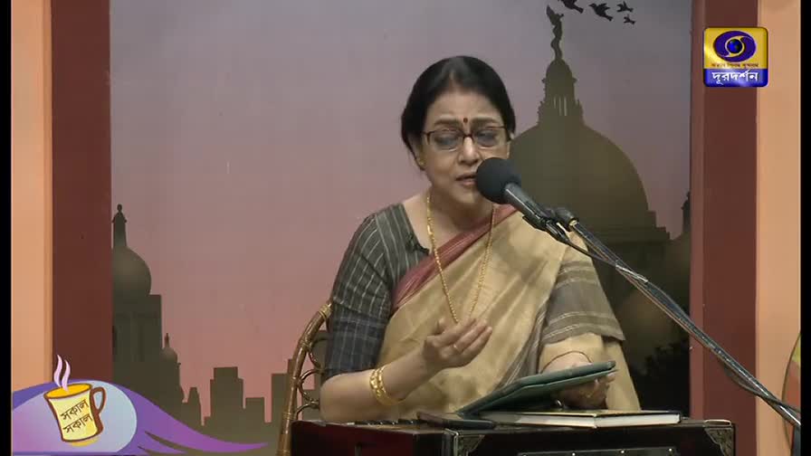 A woman in a sari sings into a microphone, her hand gesturing gently. The DD Bangla logo is visible in the corner, and a cityscape backdrop suggests the program's setting.
