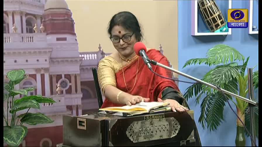 A woman in a red sari sits behind a harmonium, her hands moving over the keys and a book of lyrics. She's on a DD Bangla set, with a backdrop of a building and a logo in the corner.
