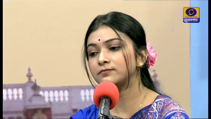 A woman with a pink flower in her hair sits before a microphone, looking down. The DD Bangla logo is visible in the corner.
A woman with a pink flower in her hair sits before a microphone, looking down. The DD Bangla logo is visible in the corner.