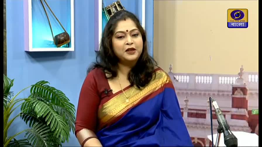 A woman in a vibrant blue and gold sari speaks into a microphone, framed by the DD Bangla logo. The set behind her features a musical instrument and a potted plant.
A woman in a vibrant blue and gold sari speaks into a microphone, framed by the DD Bangla logo. The set behind her features a musical instrument and a potted plant.